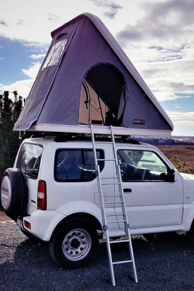 A white SUV with a rooftop tent and ladder is parked outdoors under a cloudy sky.
