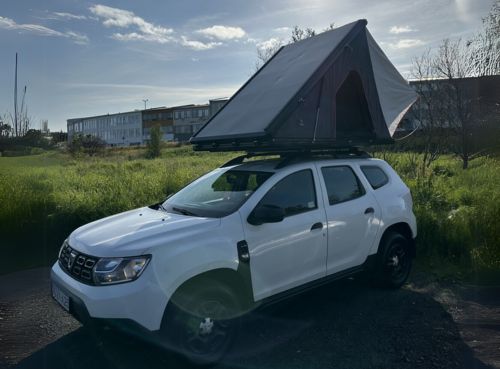 A white SUV is parked outdoors with a rooftop tent set up on top. Green grass and a building are visible in the background under a partly cloudy sky.