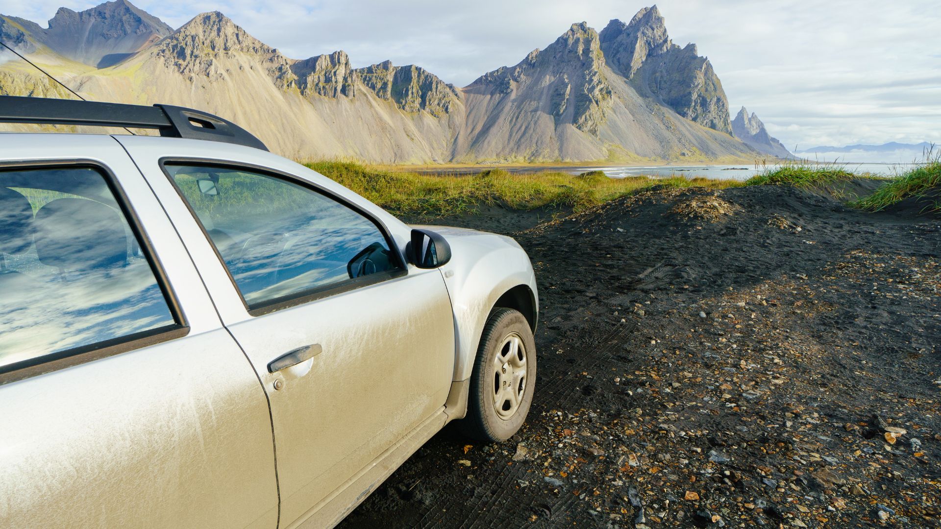 A white SUV parked on a gravel and black sand road, with tall, rugged mountains and grass in the background under a partly cloudy sky.