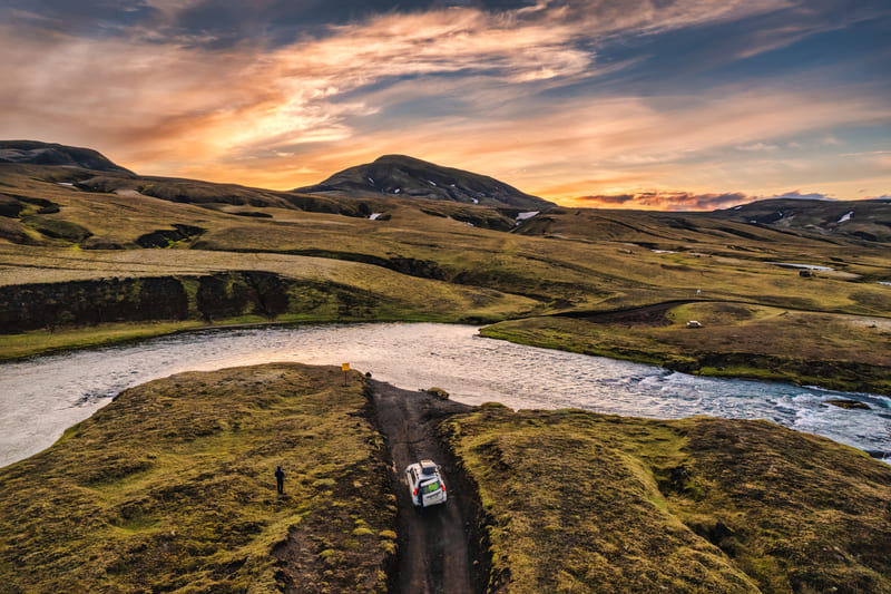 A car drives on a narrow dirt road beside a winding river in a grassy, hilly landscape at sunset with mountains in the background.