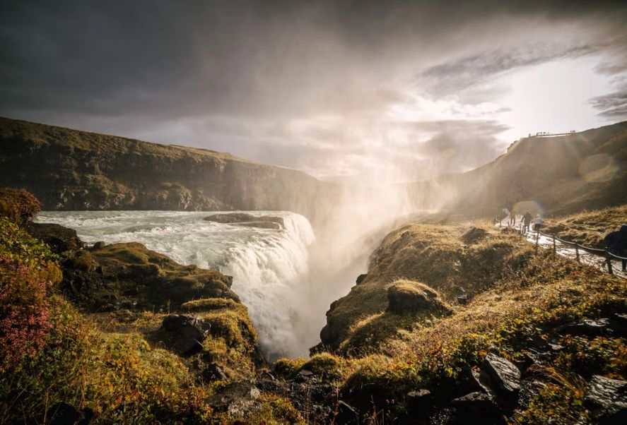 A wide waterfall flows between rocky, grassy cliffs under a cloudy sky, with mist rising and several people walking along a nearby path.
