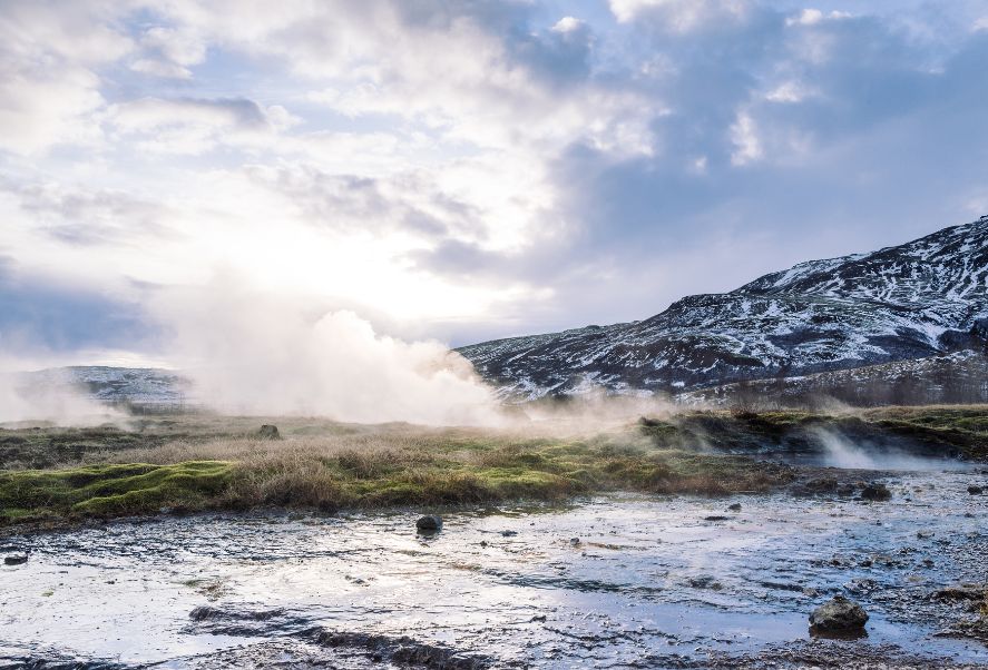 A geothermal area with steam rising from the ground, surrounded by mossy terrain and snow-covered hills under a cloudy sky.