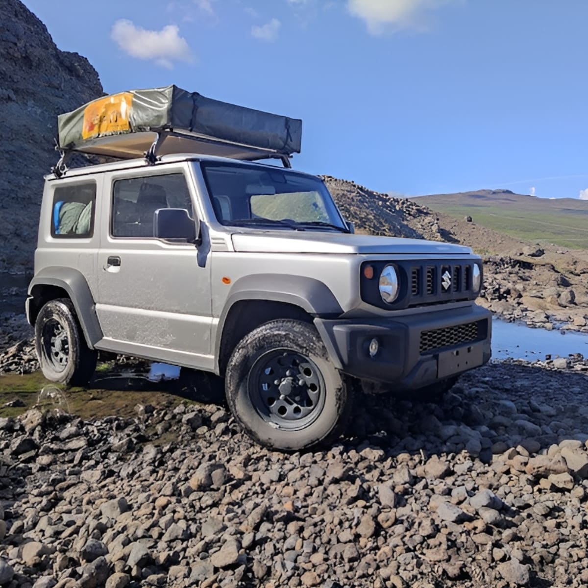 A silver off-road vehicle with a roof tent is parked on rocky terrain near a small stream and hills under a clear sky.