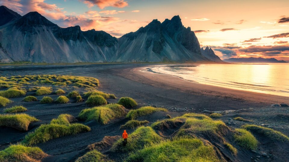 A person in an orange jacket stands among grassy mounds on a black sand beach with mountains and a bright sunset in the background.