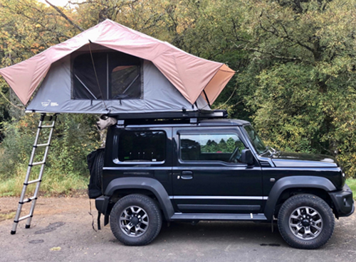 A black SUV with a rooftop tent set up and a ladder leading to the tent, parked on a road beside trees.