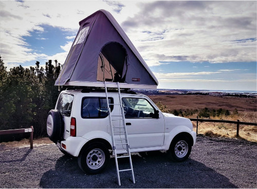 A white SUV is parked outdoors with a rooftop tent set up and a ladder attached, under a partly cloudy sky near open fields.