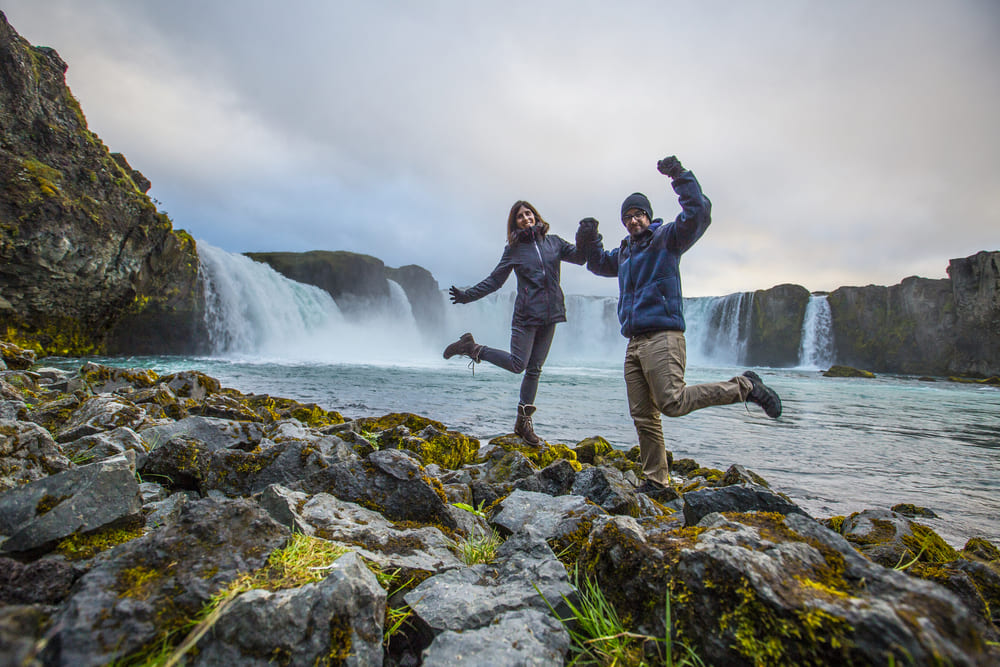 Two people in outdoor clothing jump and pose on rocky terrain near a large waterfall, with water and cliffs in the background under a cloudy sky.