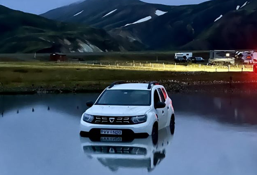 A white SUV is partially submerged in water in a remote, mountainous landscape at dusk, with a few vehicles and campers in the background.